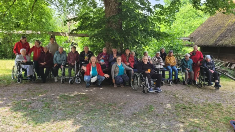 Gruppe von Senior:innen und Pflegekräften der ADAMO Tagespflege sitzt und steht gemeinsam unter einem schattigen Baum.