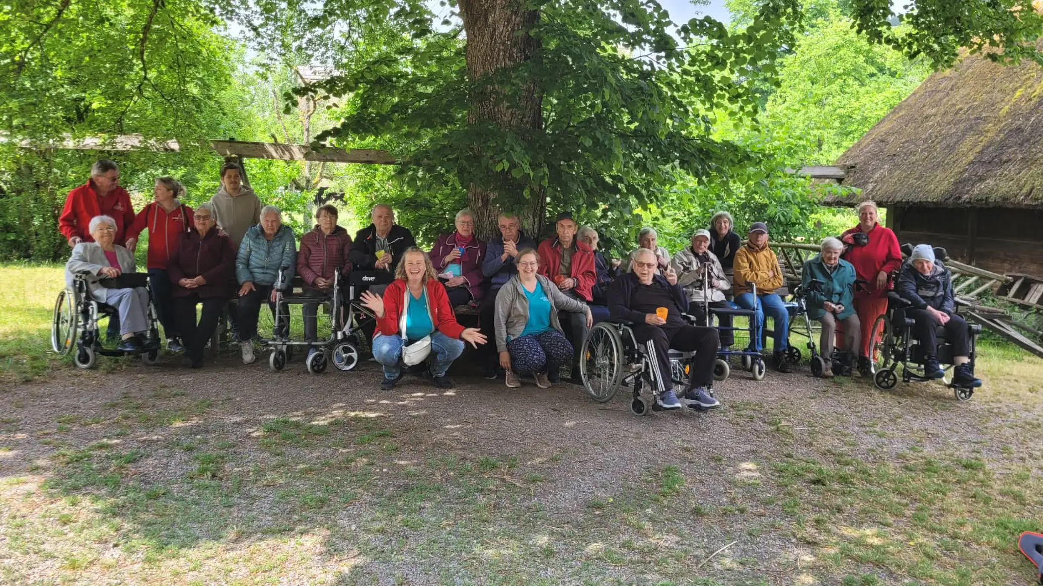 Gruppe von Senior:innen und Pflegekräften der ADAMO Tagespflege sitzt und steht gemeinsam unter einem schattigen Baum.