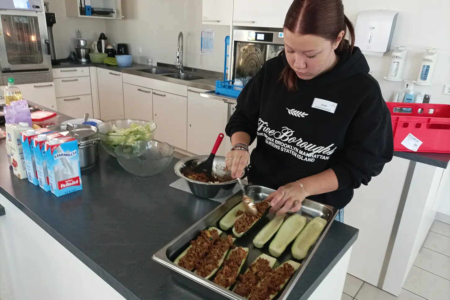 Eine junge Frau füllt die vorbereiteten Zucchinihälften mit Hackfleischmasse in einer großen Küche.