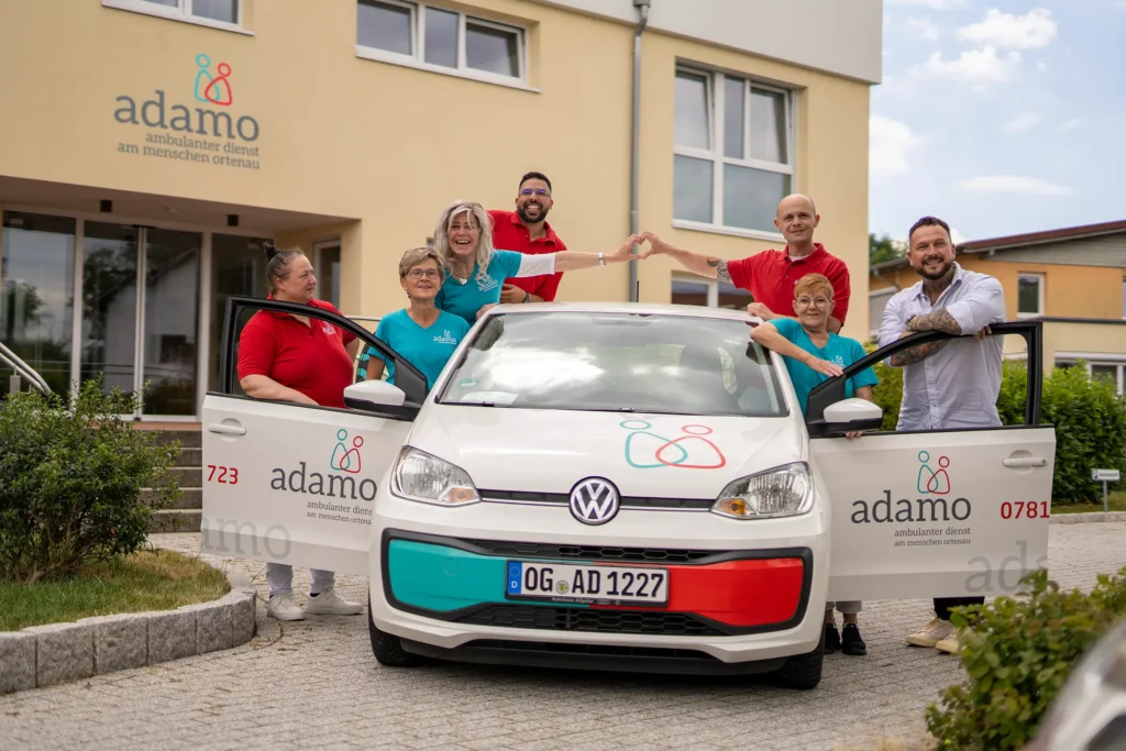 Team des ambulanten Dienstes adamo steht vor dem Gebäude in Willstätt, formt ein Herz und präsentiert ein gebrandetes Dienstfahrzeug.