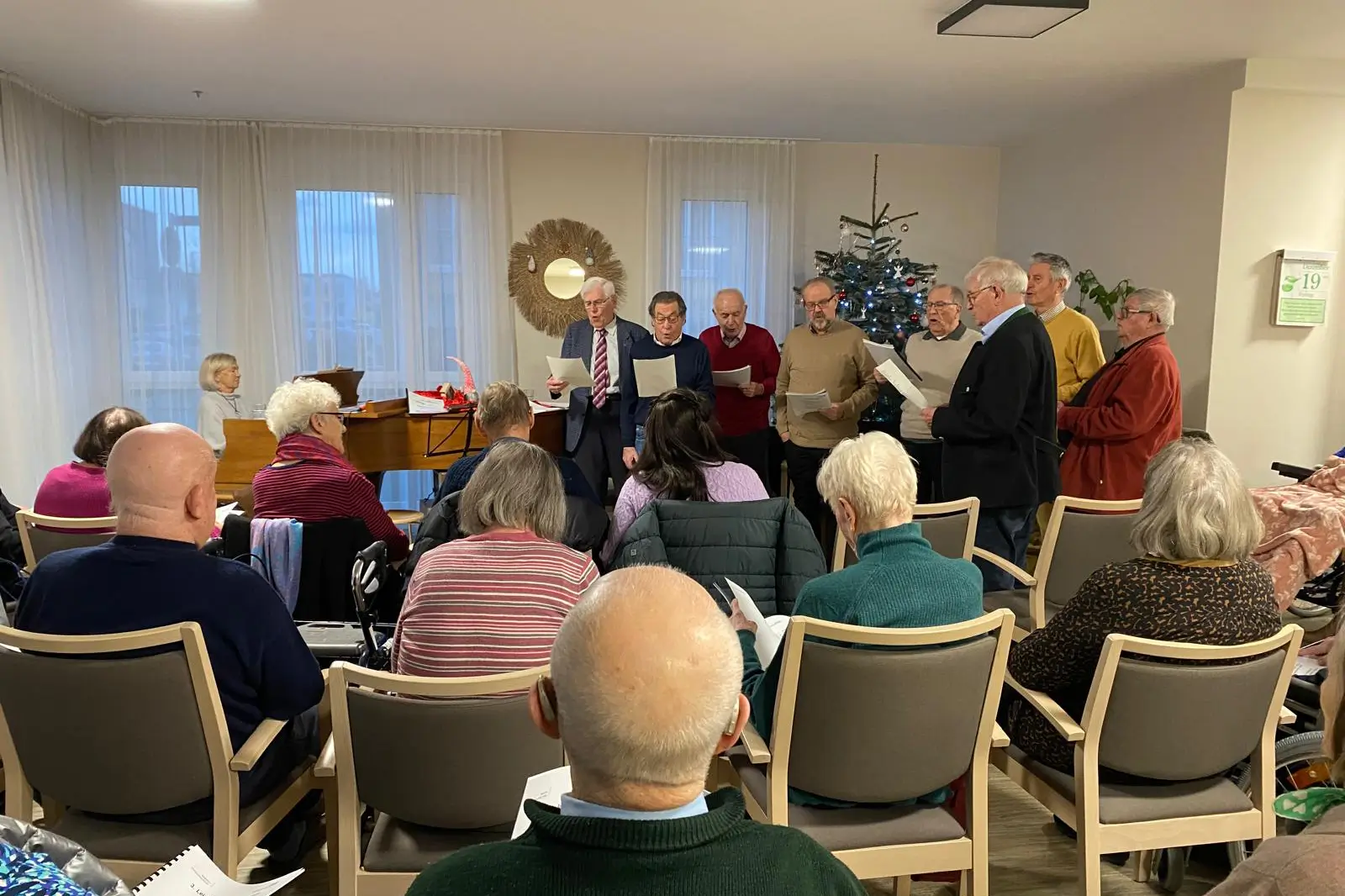 Eine Gruppe Männer singt gemeinsam Weihnachtslieder vor den Bewohnern des Seniorenzentrums, begleitet vom Klavier.