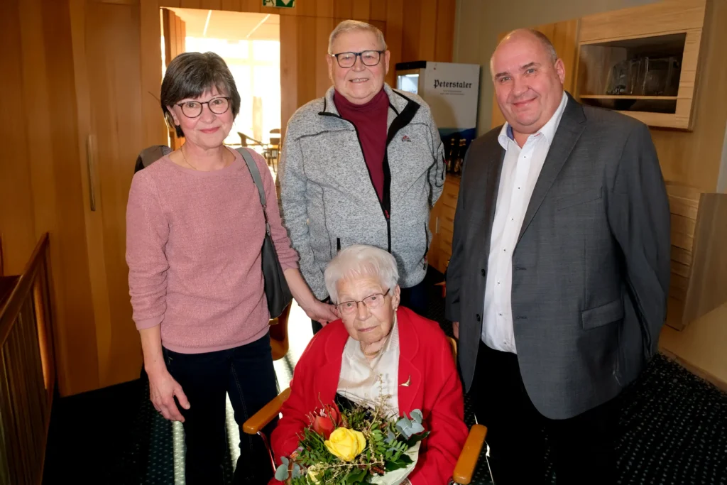 Elisabeth Huber an ihrem 100. Geburtstag im Rollstuhl mit Blumenstrauß, flankiert von zwei Gratulanten in einem festlichen Innenraum.
