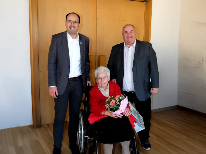Elisabeth Huber anlässlich ihres 100. Geburtstags mit Blumenstrauß, umgeben von Gratulanten bei einer Gratulation im Innenraum.