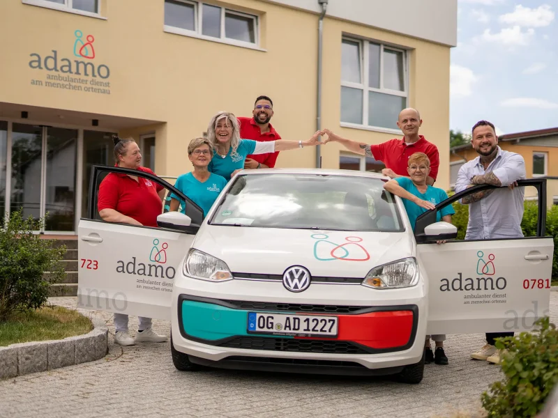 Team des ambulanten Dienstes adamo steht vor dem Gebäude in Willstätt, formt ein Herz und präsentiert ein gebrandetes Dienstfahrzeug.