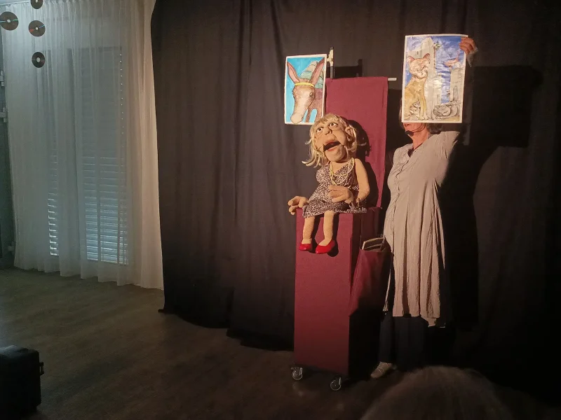 Eine Puppenspielerin steht auf der Bühne mit der Puppe Elfriede und zeigt ein Bild von Tieren.