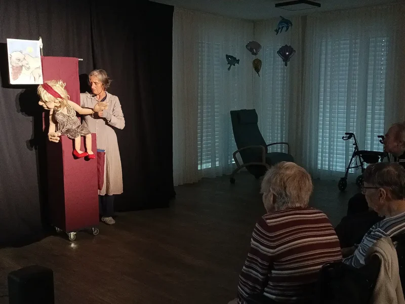 Bewohnerinnen und Bewohner verfolgen gespannt die Vorstellung mit der Figur Elfriede auf der Bühne.