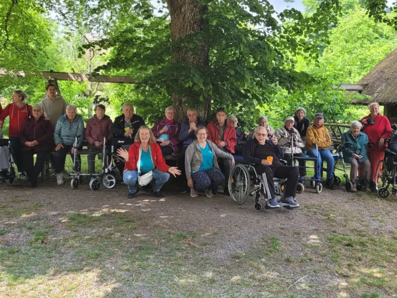 Gruppe von Senior:innen und Pflegekräften der ADAMO Tagespflege sitzt und steht gemeinsam unter einem schattigen Baum.