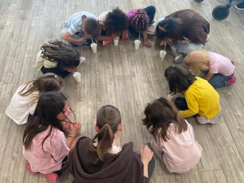 Kinder sitzen im Kreis auf dem Boden und blubbern mit Strohhalmen in Wasserbechern die Melodie von „Happy Birthday“.