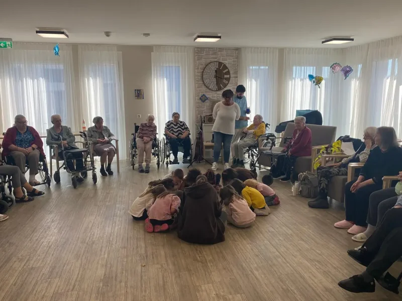 Kinder und Seniorinnen sitzen zusammen im Raum, während die Kita-Gruppe musiziert und tanzt.