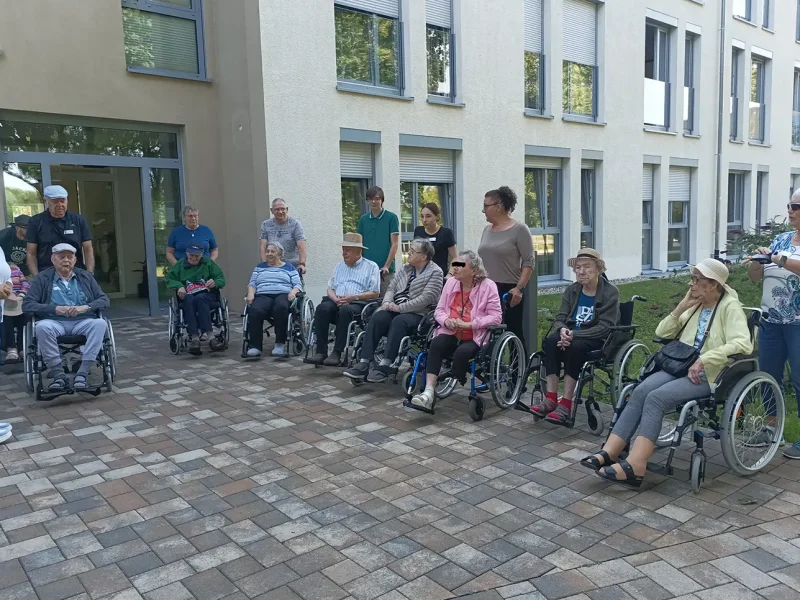 Eine Gruppe von Bewohnerinnen und Bewohnern in Rollstühlen mit Begleitpersonen versammelt sich vor dem Seniorenzentrum zum Spaziergang.