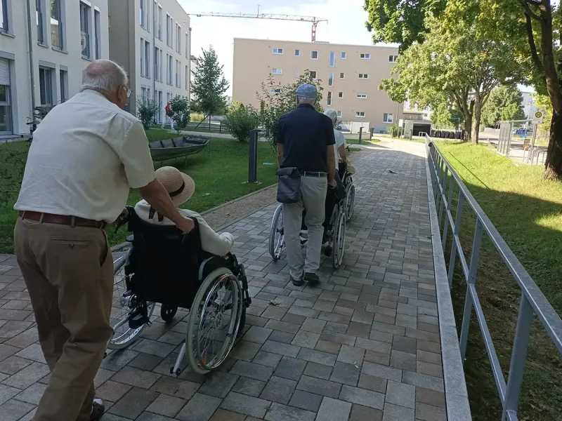Zwei Männer schieben Rollstühle auf einem Weg entlang des Seniorenzentrums in Richtung Altrhein.