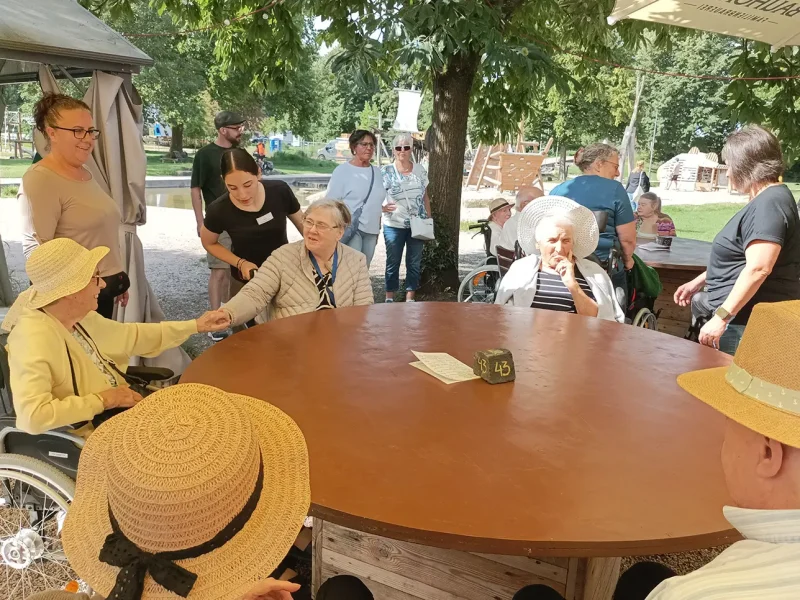Bewohnerinnen und Bewohner sitzen unter Bäumen am runden Tisch auf der Terrasse des Restaurants Rheinschnecke.