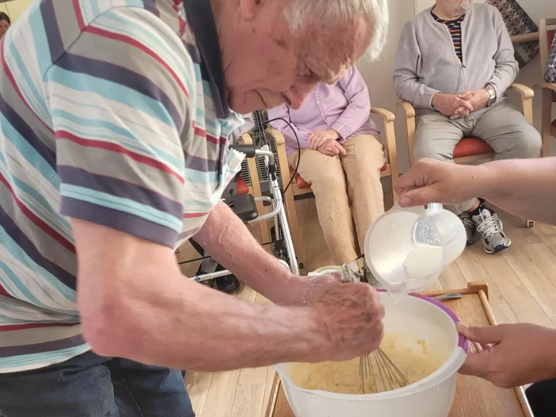 Älterer Mann rührt mit einem Schneebesen Teig in einer großen Schüssel an, während jemand Milch hinzufügt.