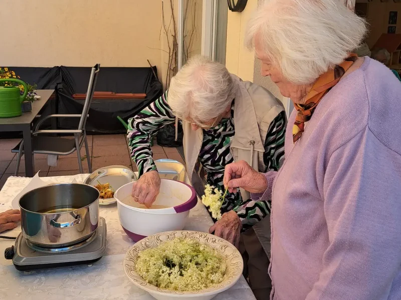 Zwei ältere Damen stehen an einem Tisch, tauchen Holunderblüten in Teig und bereiten sie fürs Ausbacken vor.