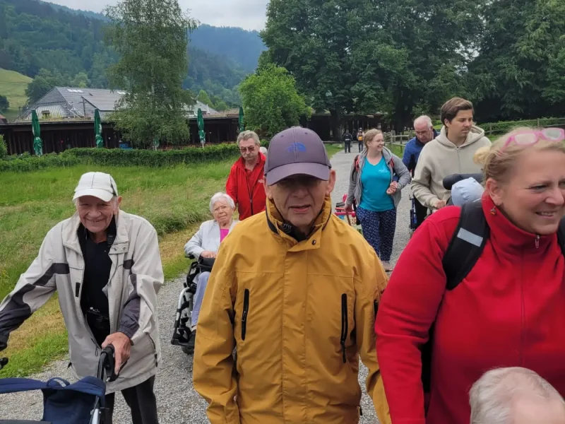 Senior:innen mit Rollatoren und Pflegekräften gehen gemeinsam einen Weg entlang, umgeben von Natur.