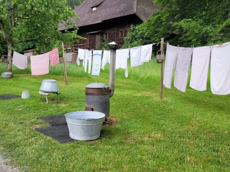 Historische Szene mit aufgehängter Wäsche, Zinkwannen und Waschofen im Garten eines alten Bauernhofs.