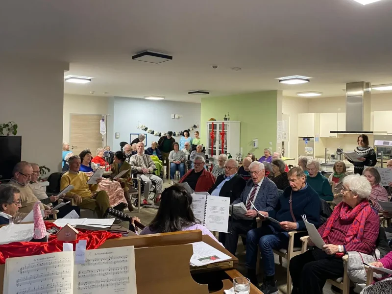 Viele Bewohner und Angehörige des Seniorenzentrums sitzen zusammen im Gemeinschaftsraum und singen gemeinsam Weihnachtslieder.