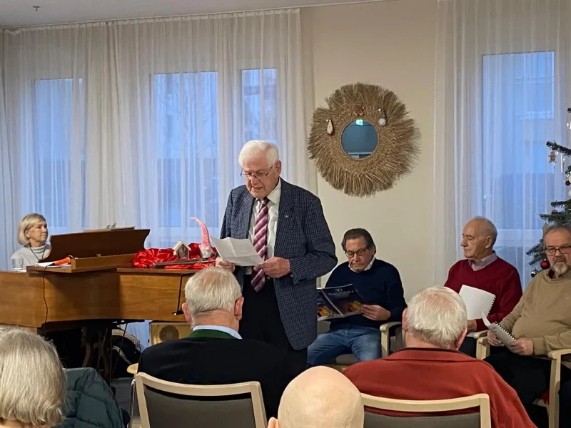 Ein älterer Herr trägt beim Weihnachtsliedernachmittag im Seniorenzentrum eine Weihnachtsgeschichte vor, während die Bewohner aufmerksam zuhören.