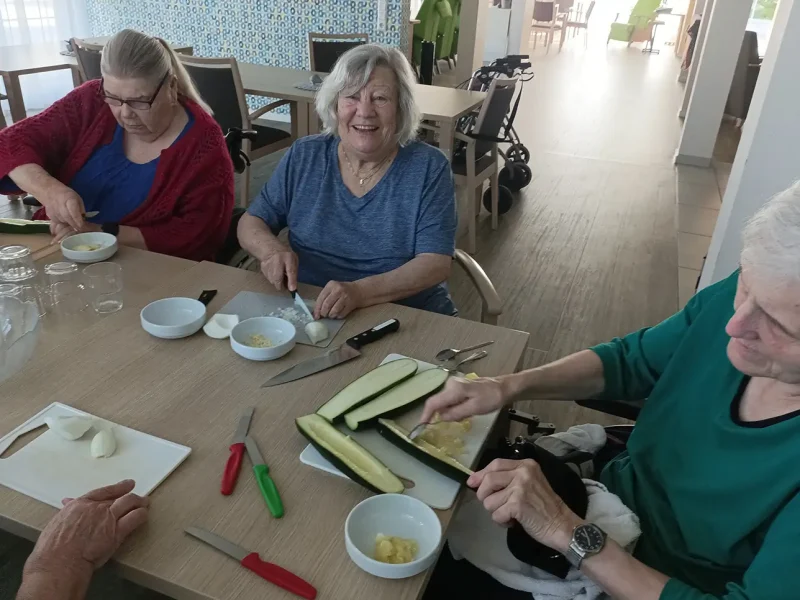 Drei Bewohnerinnen sitzen lachend am Tisch und schneiden Gemüse für das Gericht.
