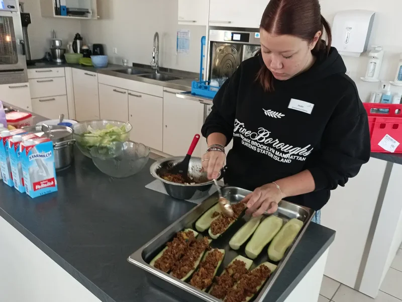 Eine junge Frau füllt die vorbereiteten Zucchinihälften mit Hackfleischmasse in einer großen Küche.
