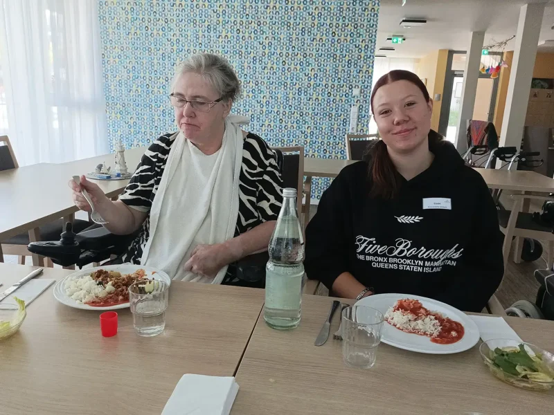 Eine Bewohnerin und eine Schülerin sitzen beim Essen am Tisch und lächeln zufrieden.