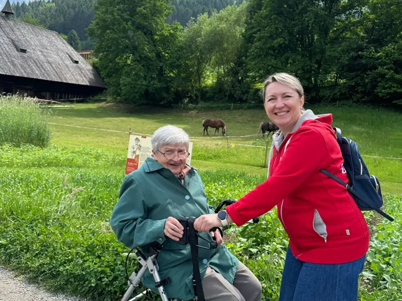 Seniorin mit Rollator und ADAMO-Mitarbeiterin lächeln beim Spaziergang vor grüner Wiese und Fachwerkgebäuden.