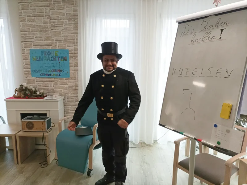 Schornsteinfeger steht vor mehreren Bewohnerinnen und Bewohnern im Aufenthaltsraum des Seniorenzentrums.